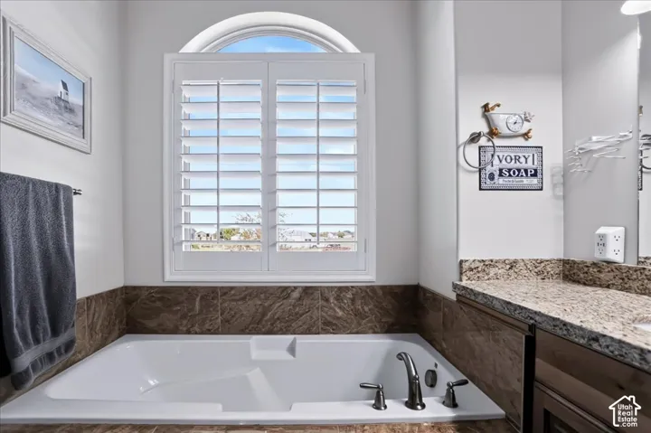 Built-in soaking tub with dark stone-finish surround, white plantation shutters, arched transom window, and stone-finish vanity countertop