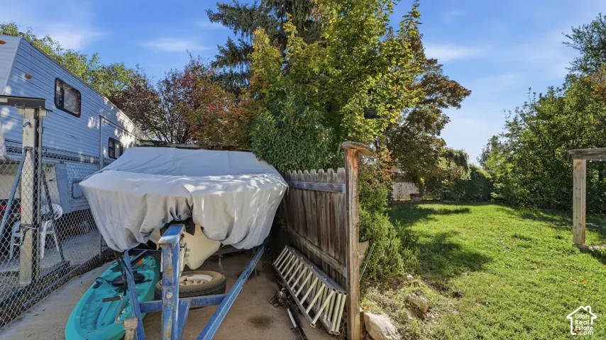 Backyard featuring a paved side parking area, wood-slat fencing, mature tree canopy, and open grassy space
