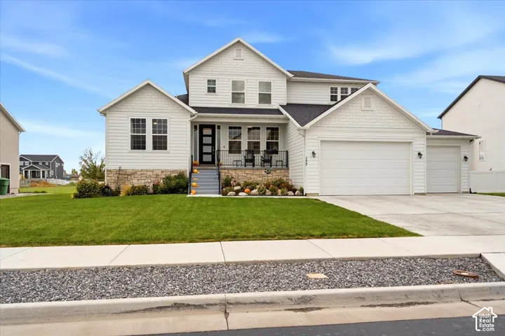 Two-story exterior featuring white horizontal lap and shingle siding, stacked stone foundation accents, a multi-gabled roofline, a covered front porch with metal railing, and an attached three-car garage
