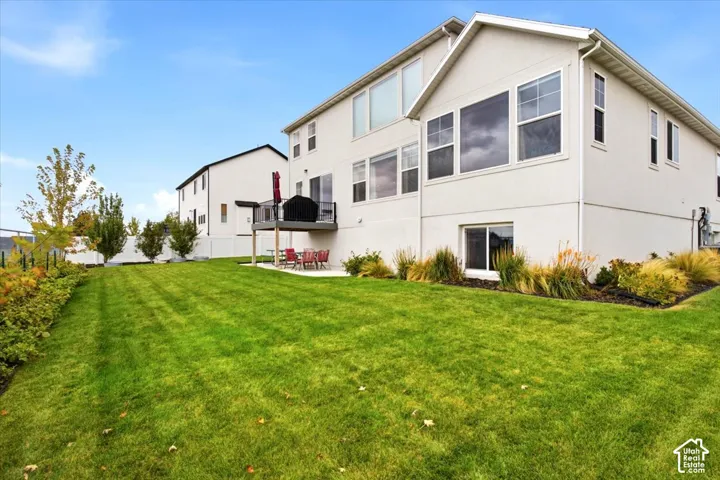 Spacious rear exterior featuring a manicured lawn, elevated deck with dark metal railing, concrete patio, white vinyl fencing, and drought-tolerant landscaping
