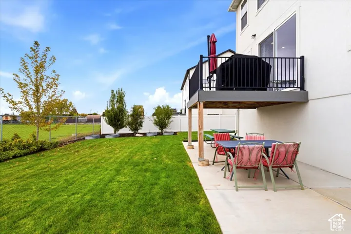 Backyard featuring a concrete patio, elevated deck with black metal railing, manicured lawn, and white vinyl perimeter fencing