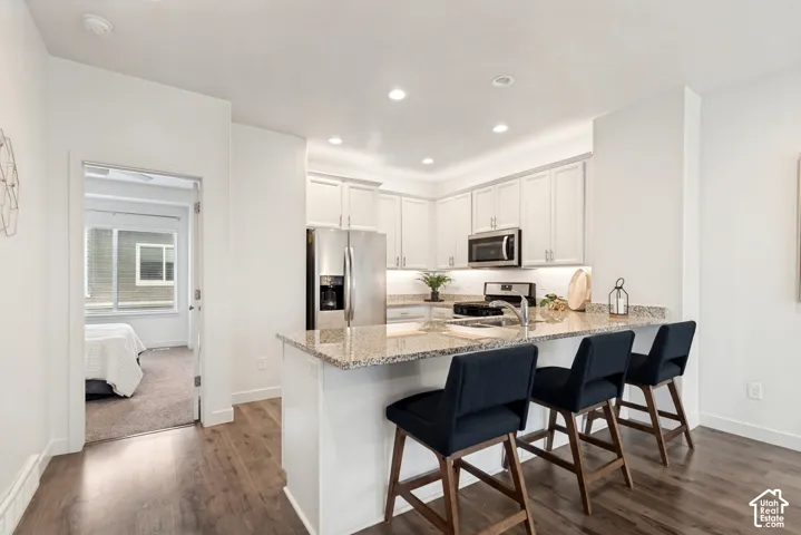 Kitchen featuring white shaker-style cabinetry, stone-surface countertops with a breakfast bar, stainless steel appliances, recessed ceiling lighting, and wood-finish flooring