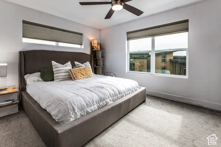 Neutral-toned bedroom featuring carpeted flooring, a ceiling fan with integrated lighting, built-in corner shelving, and dual windows equipped with pleated shades
