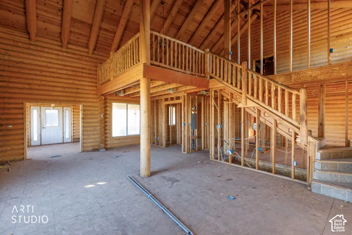 Open-concept interior featuring exposed log walls, vaulted ceilings, a structural wooden staircase, a built-in balcony railing, and natural light via oversized window apertures