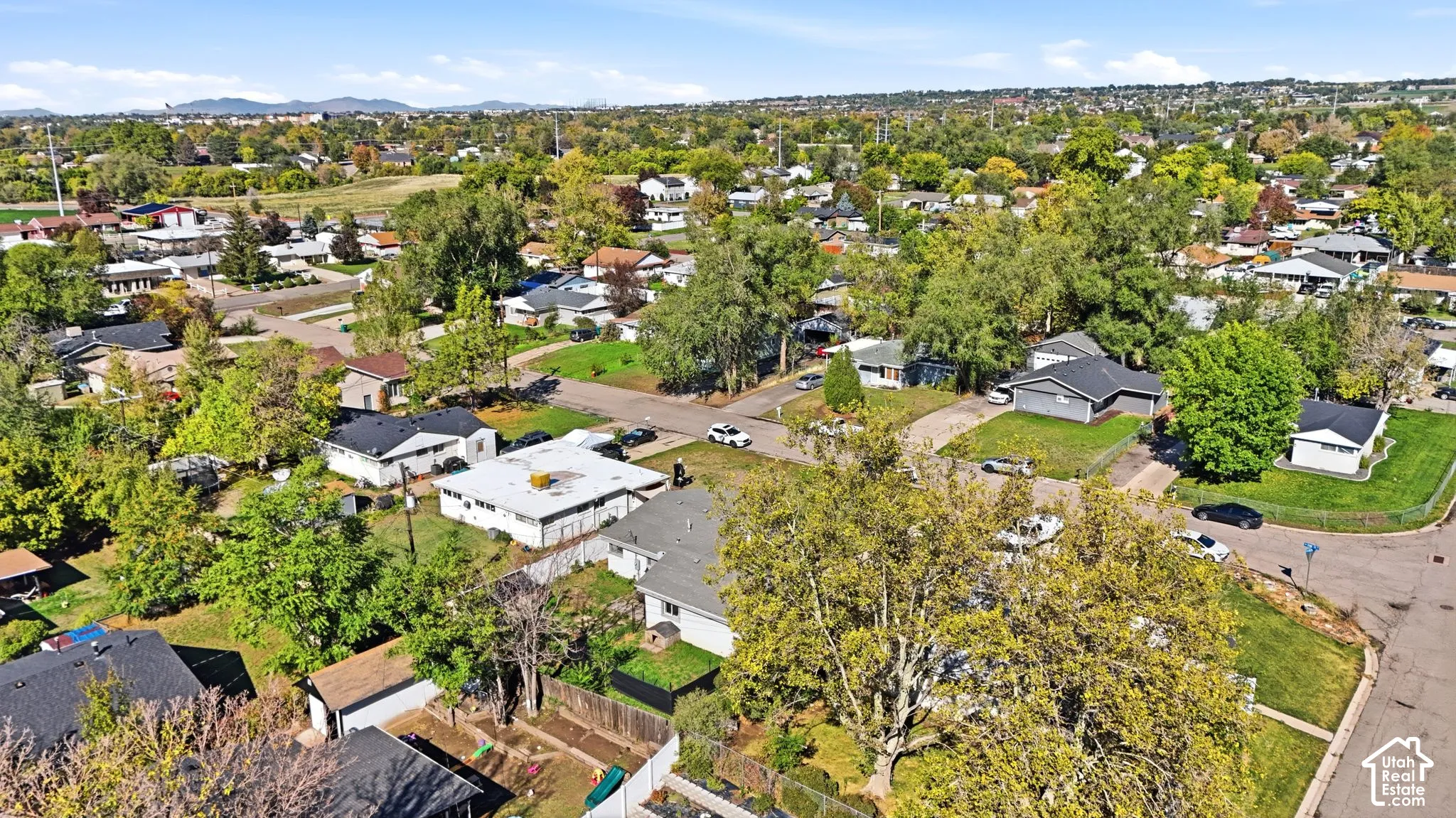 Aerial perspective of a residential setting featuring mature trees, manicured lawns, paved streets, and mountain terrain in the distance