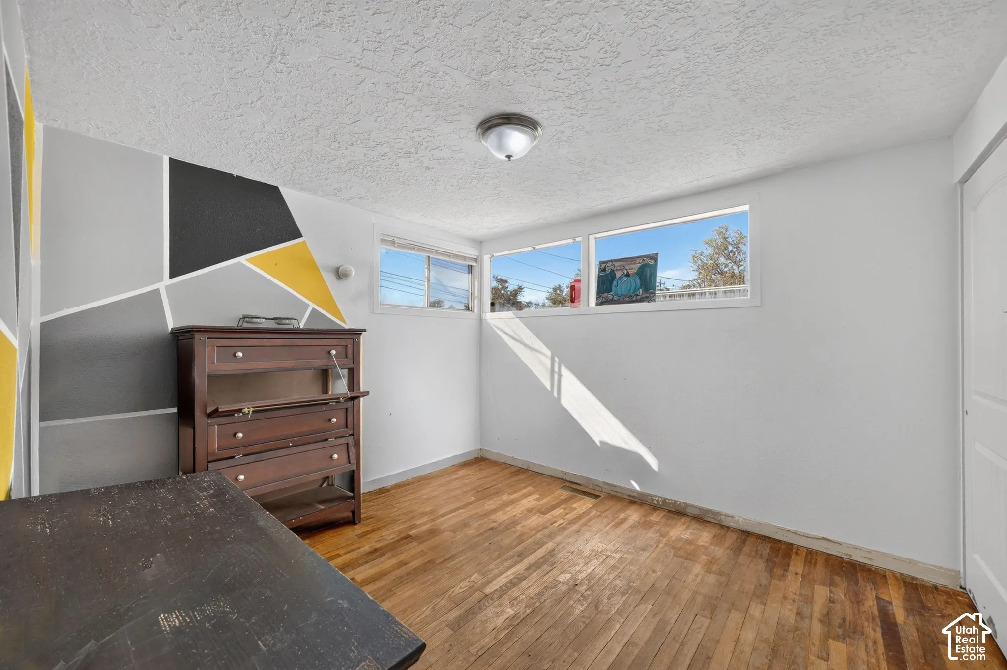 Room interior featuring wood-finish flooring, white walls with geometric accent paint, flush-mount ceiling lighting, and large windows