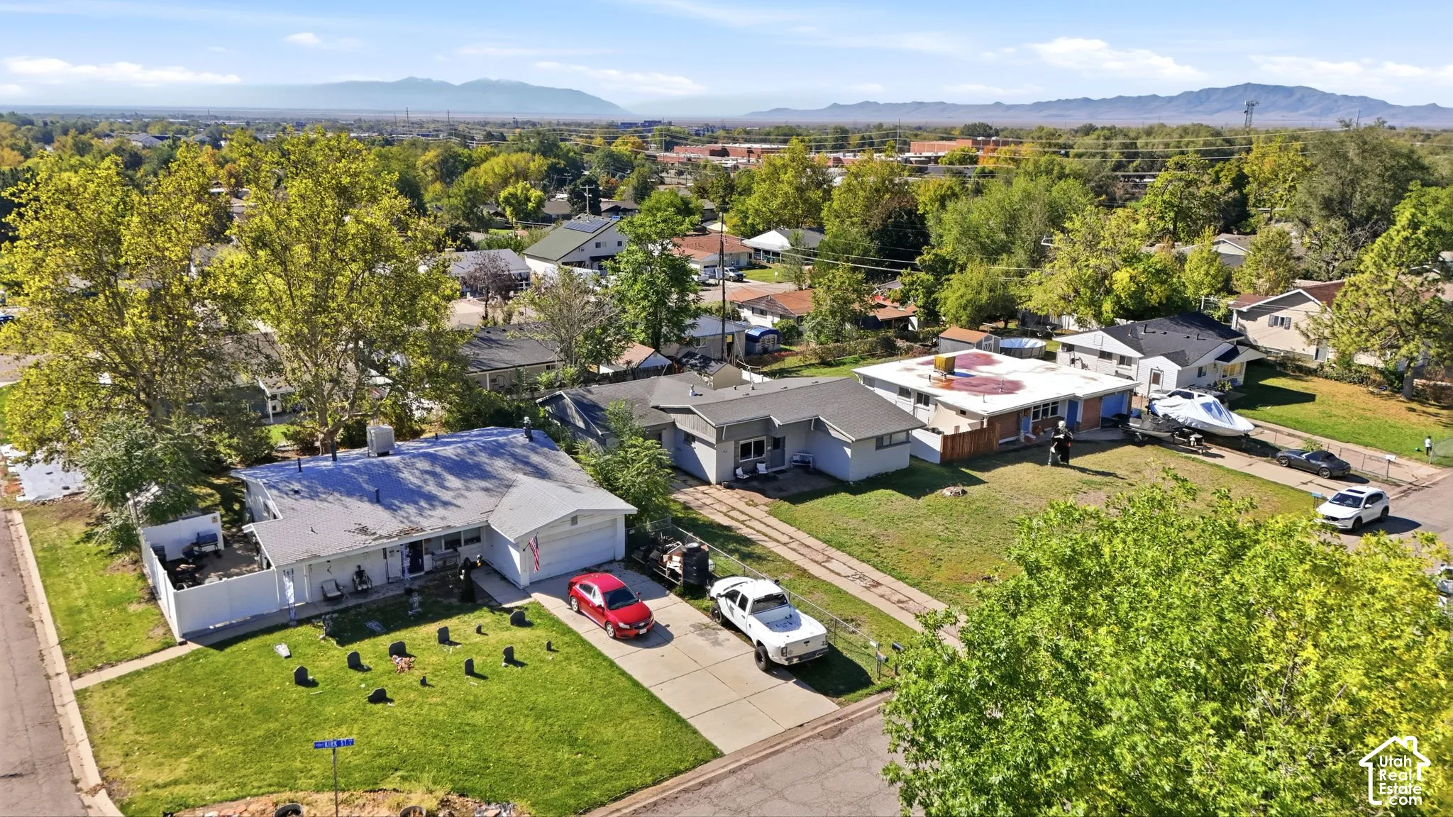 Aerial perspective of residential properties featuring attached garages, concrete driveways, and expansive lot space with mature trees set against a mountain backdrop