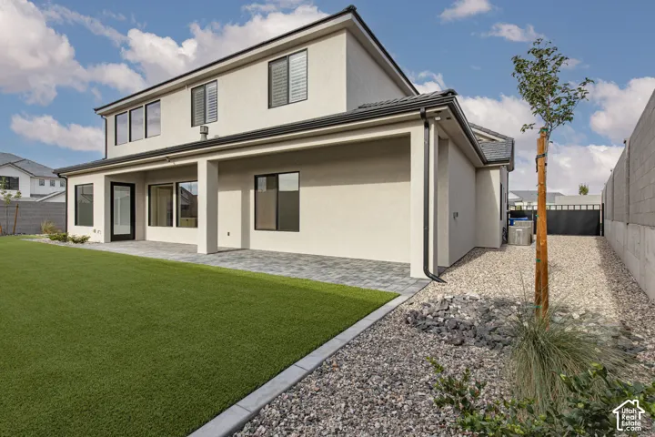 Stucco exterior featuring a covered paver patio, synthetic turf lawn, stone-accented landscape border, and perimeter masonry walls