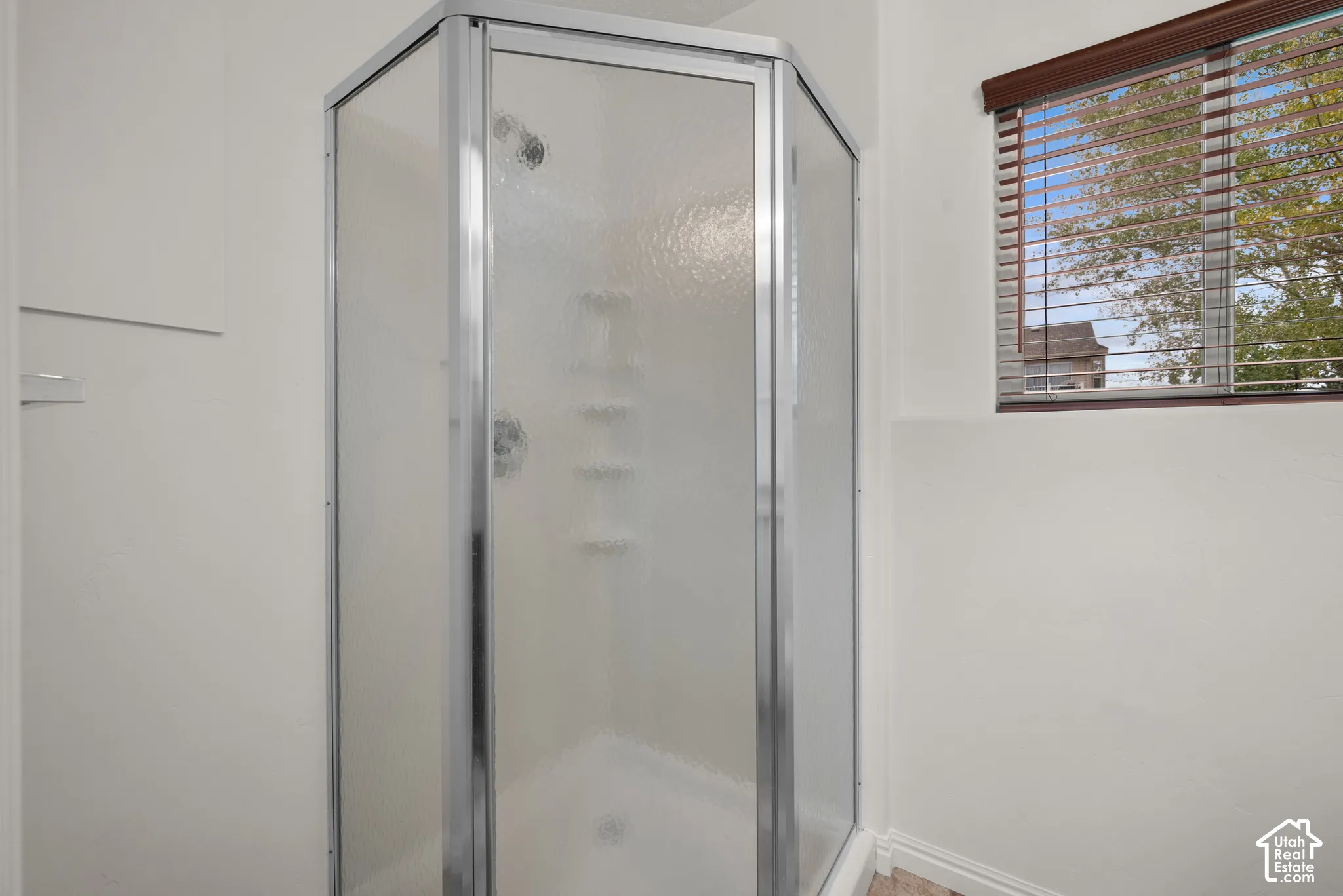 Corner shower enclosure featuring a metallic frame, textured glass panels, integrated shelving, and a horizontal window with fitted blinds