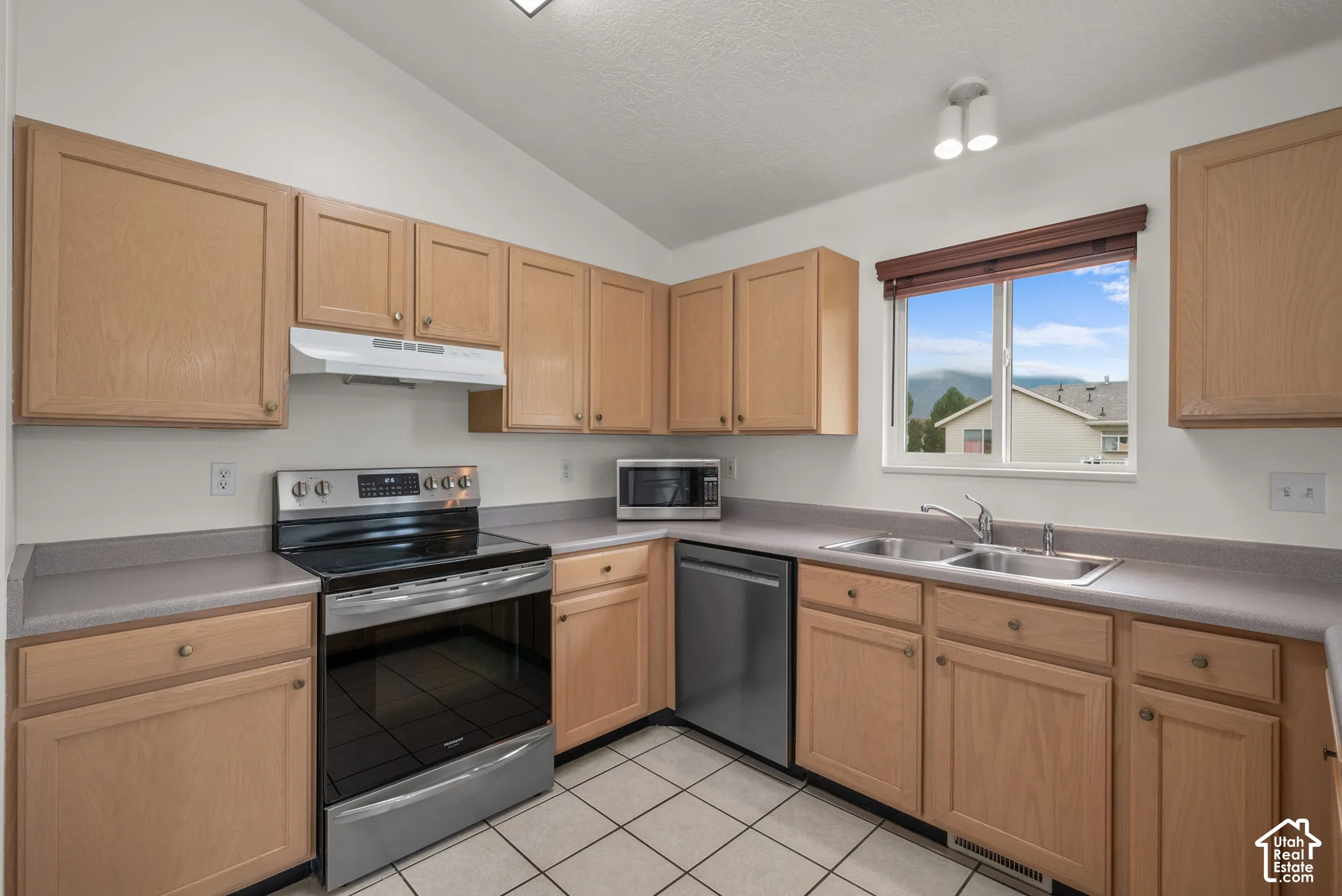 Kitchen featuring natural-finish wood cabinetry, solid-surface countertops, stainless steel appliances, tile flooring, and a double-basin sink