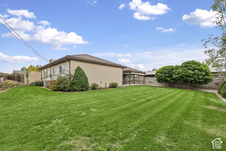 Expansive backyard featuring a manicured lawn, masonry exterior walls, a covered carport, mature trees, and perimeter privacy fencing