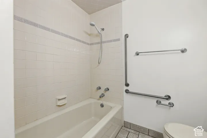 Bathtub featuring a tiled surround with decorative inlay, handheld shower head, and wall-mounted metal support bars
