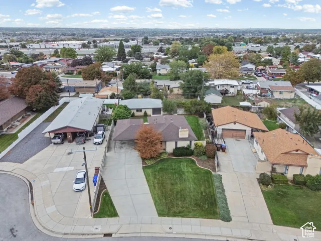 Aerial perspective featuring a single-story residence with a multi-car covered parking area, expansive concrete driveway, masonry chimney, and manicured front lawn