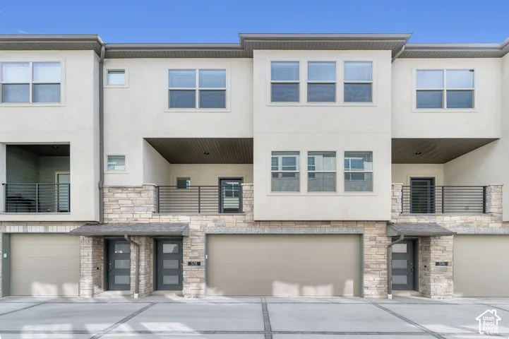 Multi-level exterior featuring light-toned stucco, natural stone veneer accents, recessed balconies with metal railings, and multi-panel entry doors