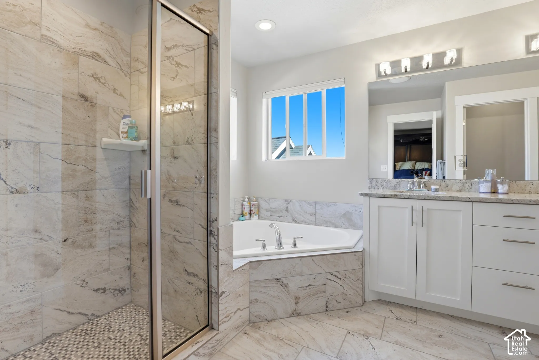 Bathroom featuring a walk-in shower with stone-finish tile surround, corner soaking tub, dual-sink vanity with stone-finish countertop, and tiled flooring