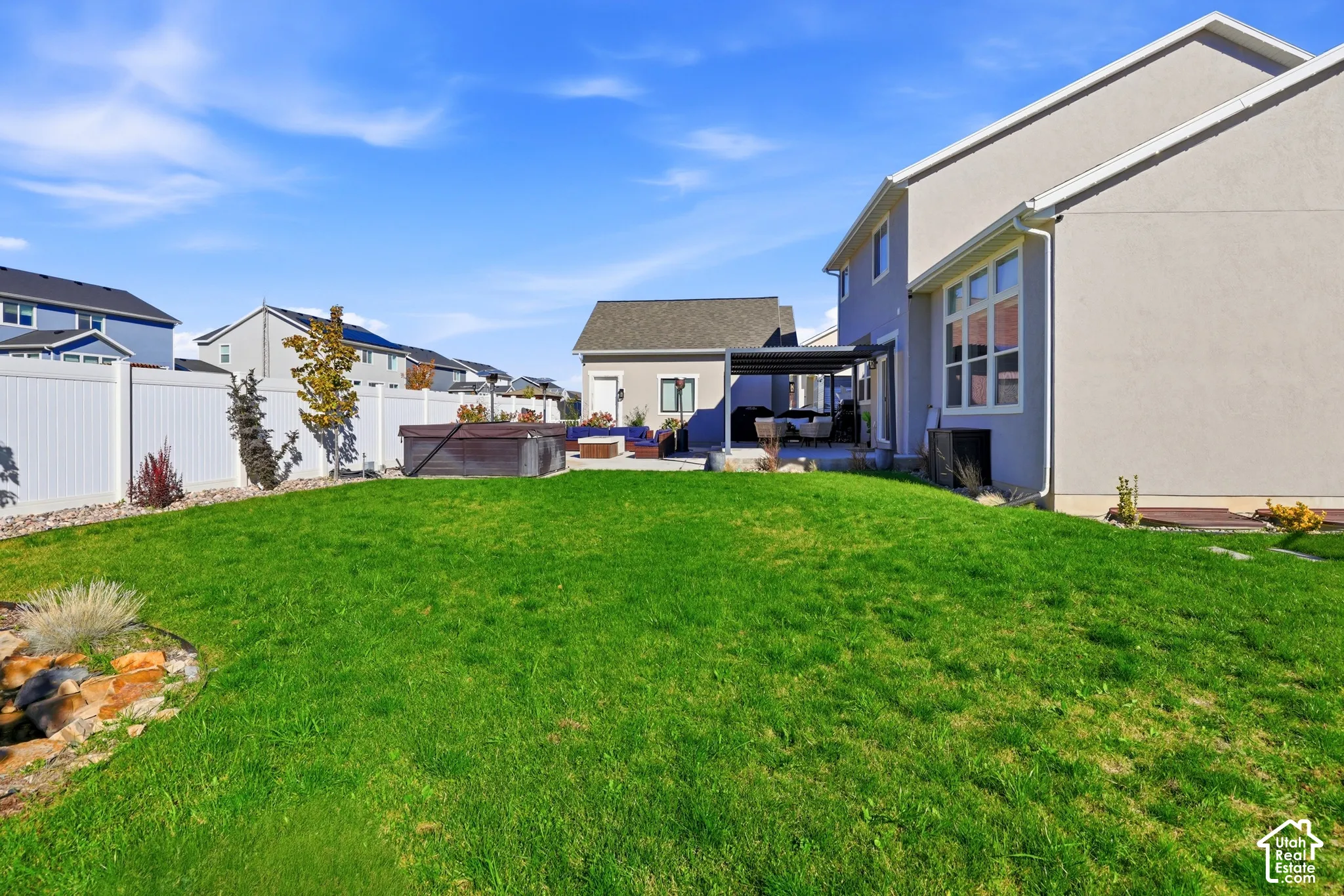 Rear exterior featuring a manicured lawn, white vinyl privacy fencing, stone hardscaping, a detached auxiliary structure, and a covered patio area