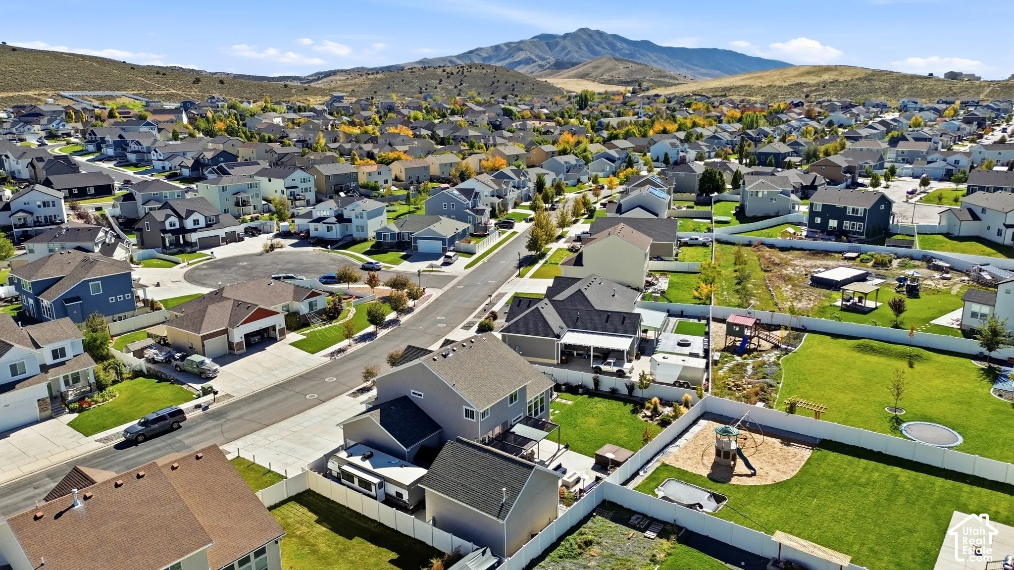 Aerial perspective of the residential subdivision featuring expansive grass lawns, white privacy fencing, asphalt paved streets, and a prominent mountain backdrop