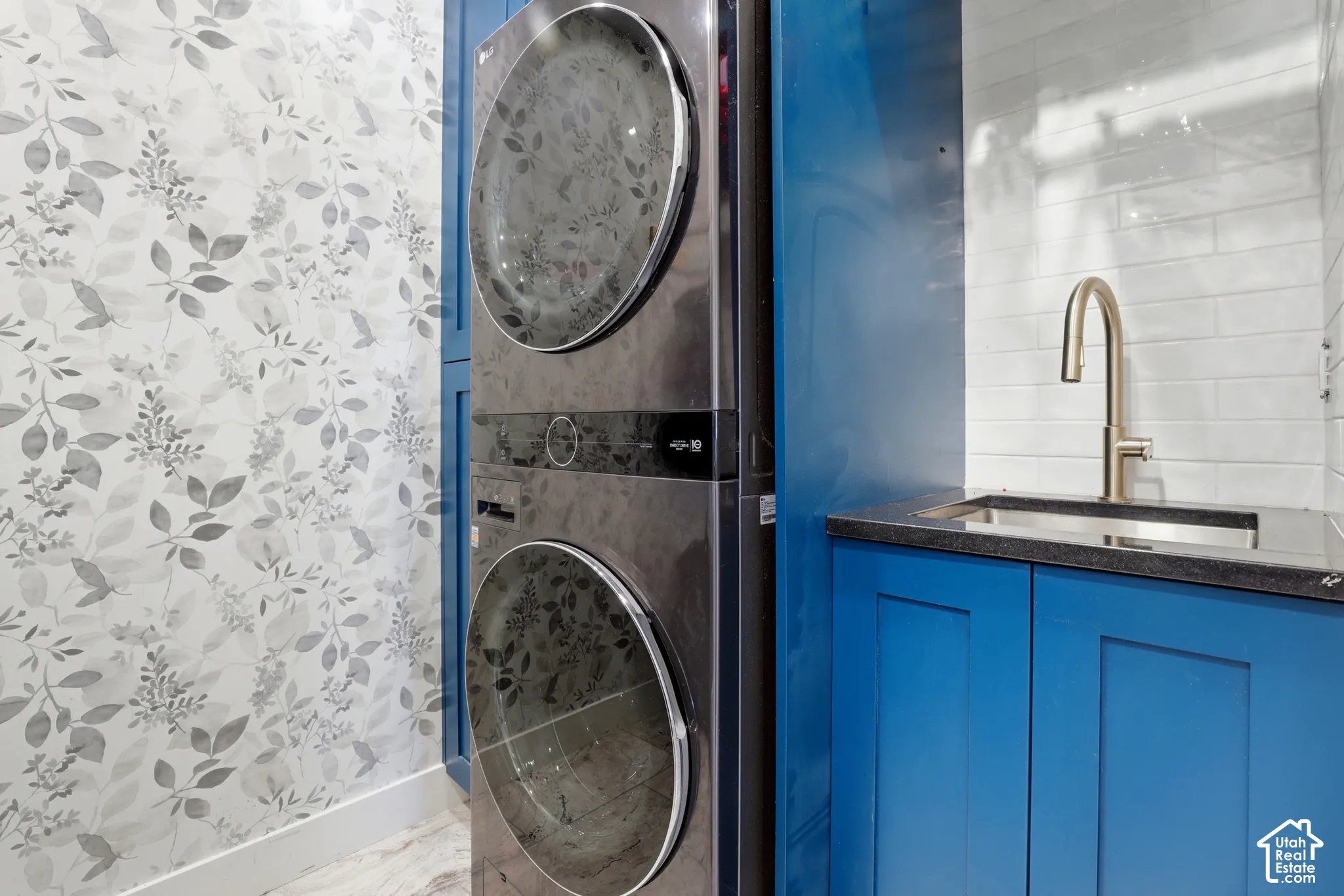 Stacked laundry unit with dark metallic finish, deep blue cabinetry, stone-surface countertop, undermount sink with arched metal faucet, and subway tile backsplash
