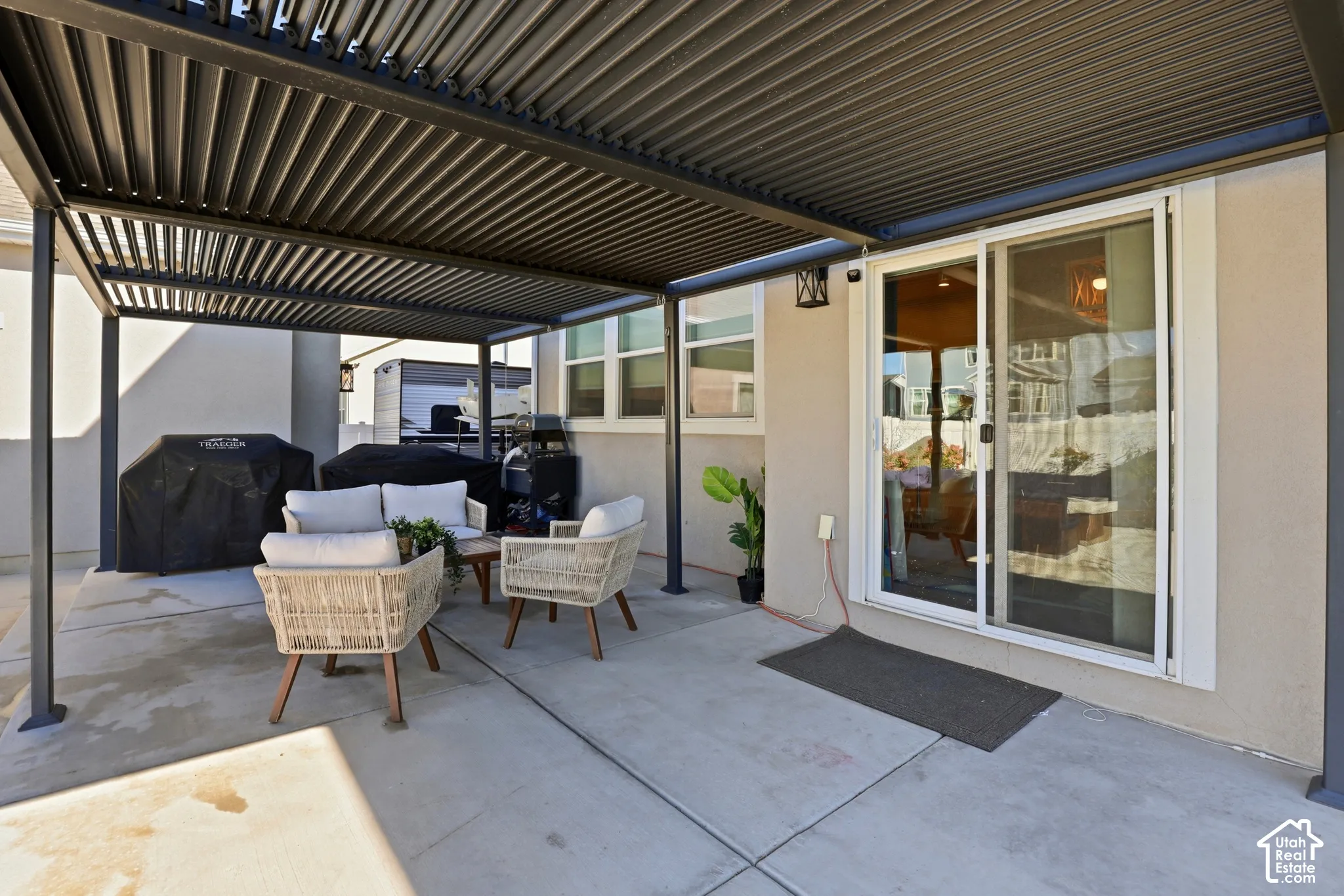 Covered concrete patio featuring a black metal louvered pergola, sliding glass door, and exterior wall-mounted lighting
