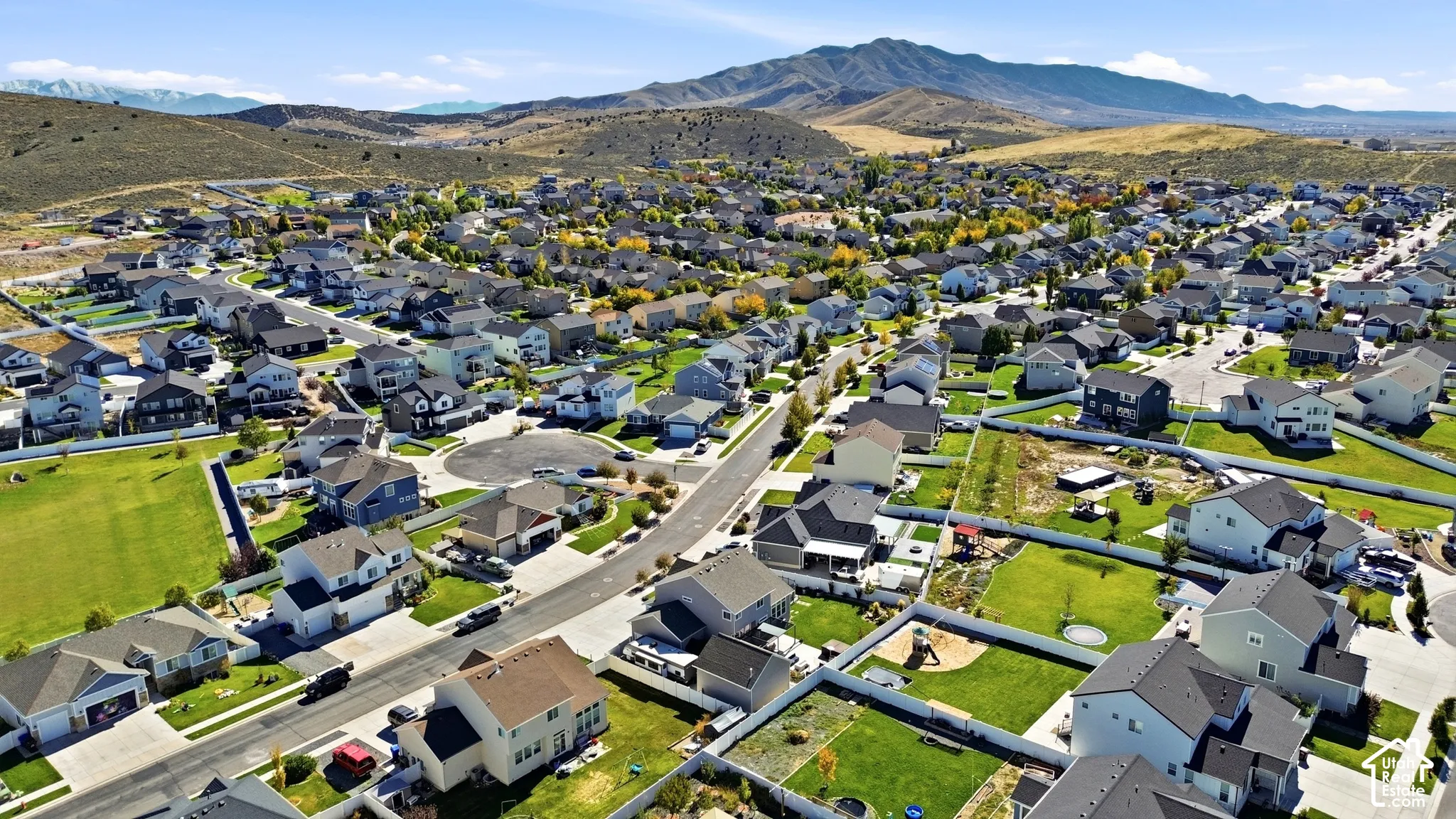 Aerial perspective of a residential neighborhood featuring asphalt paved streets, varied architectural styles, fenced residential lots, and an expansive mountain range backdrop