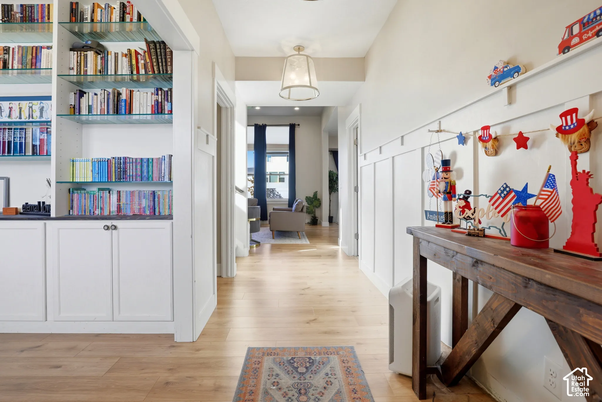 Hallway featuring wood-finish flooring, built-in cabinetry with glass shelving, wall-mounted board and batten wainscoting, and a pendant light fixture