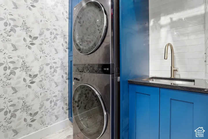 Stacked laundry unit with dark metallic finish, deep blue cabinetry, stone-surface countertop, undermount sink with arched metal faucet, and subway tile backsplash