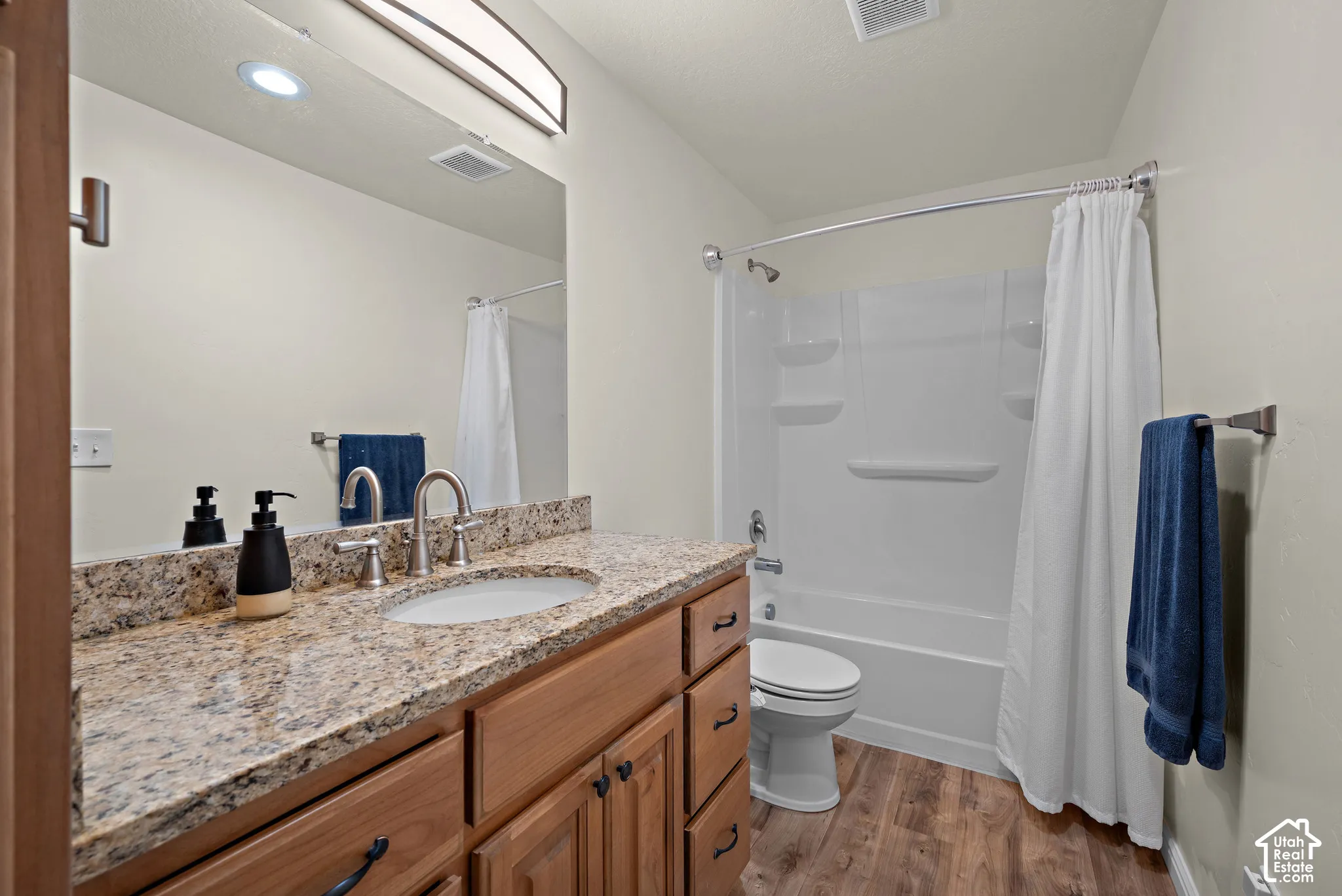 Bathroom featuring a stone-patterned vanity countertop, wood-finish cabinetry, an oval undermount sink, a polished metal faucet, and wood-finish flooring