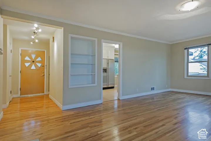 Living area featuring wood-finish flooring, built-in shelving, crown molding, and a solid-core front door with decorative glass insets