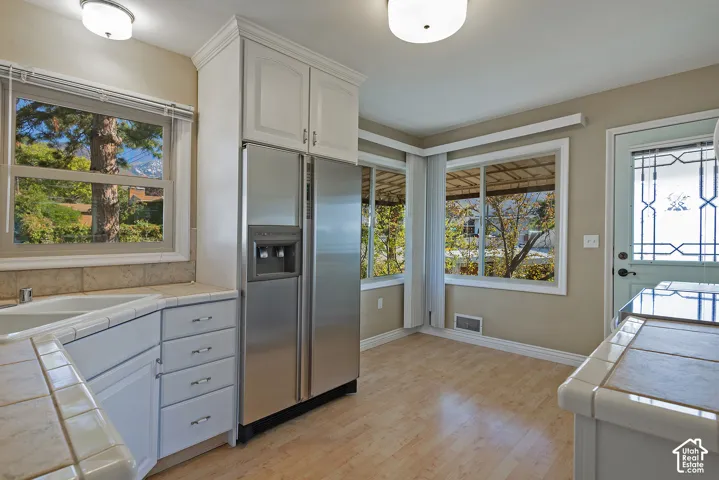 Kitchen featuring tile countertops, white cabinetry with crown molding, stainless steel side-by-side refrigerator, light-toned wood-finish flooring, and multiple windows