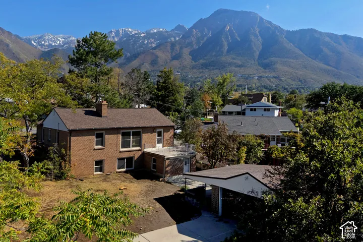 brick exterior featuring a shingle roof, chimney, concrete driveway, and attached carport against a backdrop of prominent mountain ranges