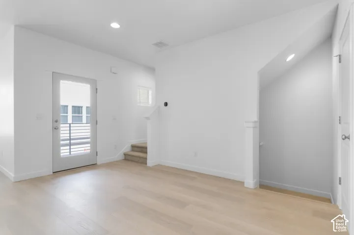 Interior space featuring light wood-finish flooring, recessed lighting, white wall-mounted trim, a half-glass exterior door, and a carpeted staircase with a partial wall
