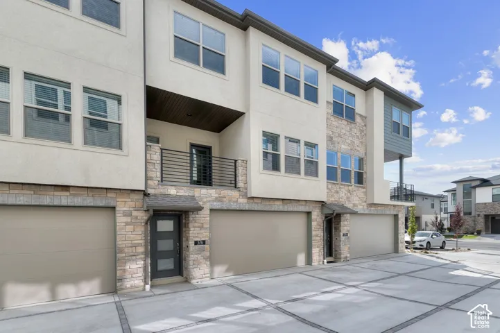 Multi-level exterior featuring light-toned stucco and stacked-stone veneer, metal balcony railing, attached multi-car garages, and a private entry with a frosted glass panel door