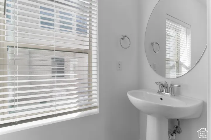Pedestal sink with a double-handle chrome faucet, oval wall-mounted mirror, and horizontal slat window blinds