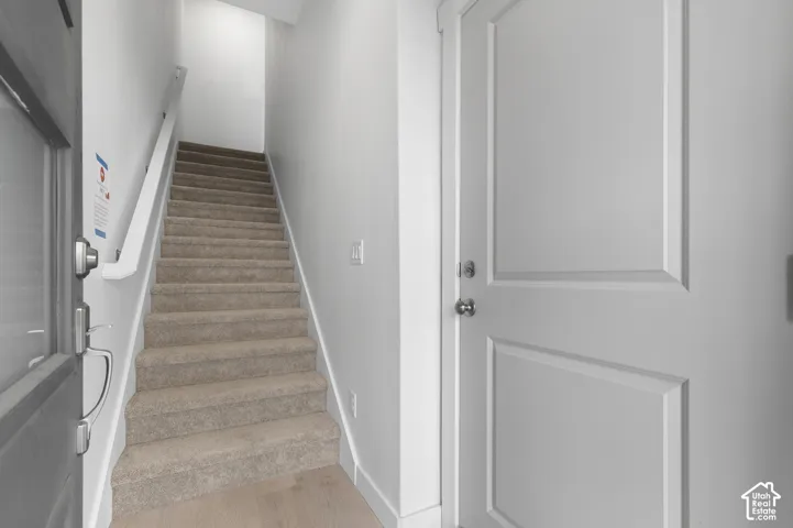 Entryway featuring a solid-core door with brushed nickel hardware, neutral carpeted staircase, light-toned wood-finish flooring, and white wall-mounted handrail