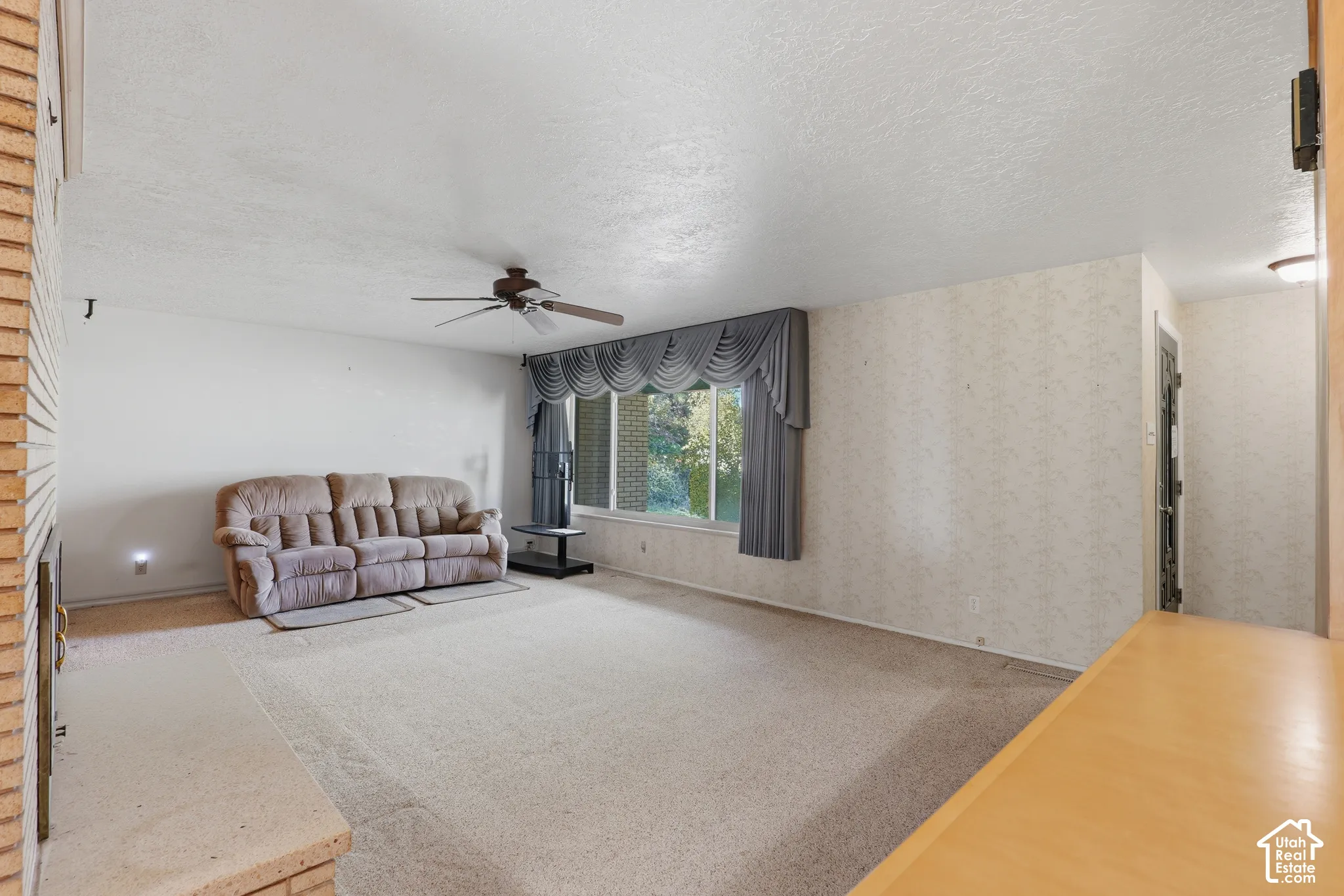 Living area featuring a textured brick fireplace surround, neutral wall-to-wall carpeting, a multi-blade ceiling fan, and large windows with integrated hardware