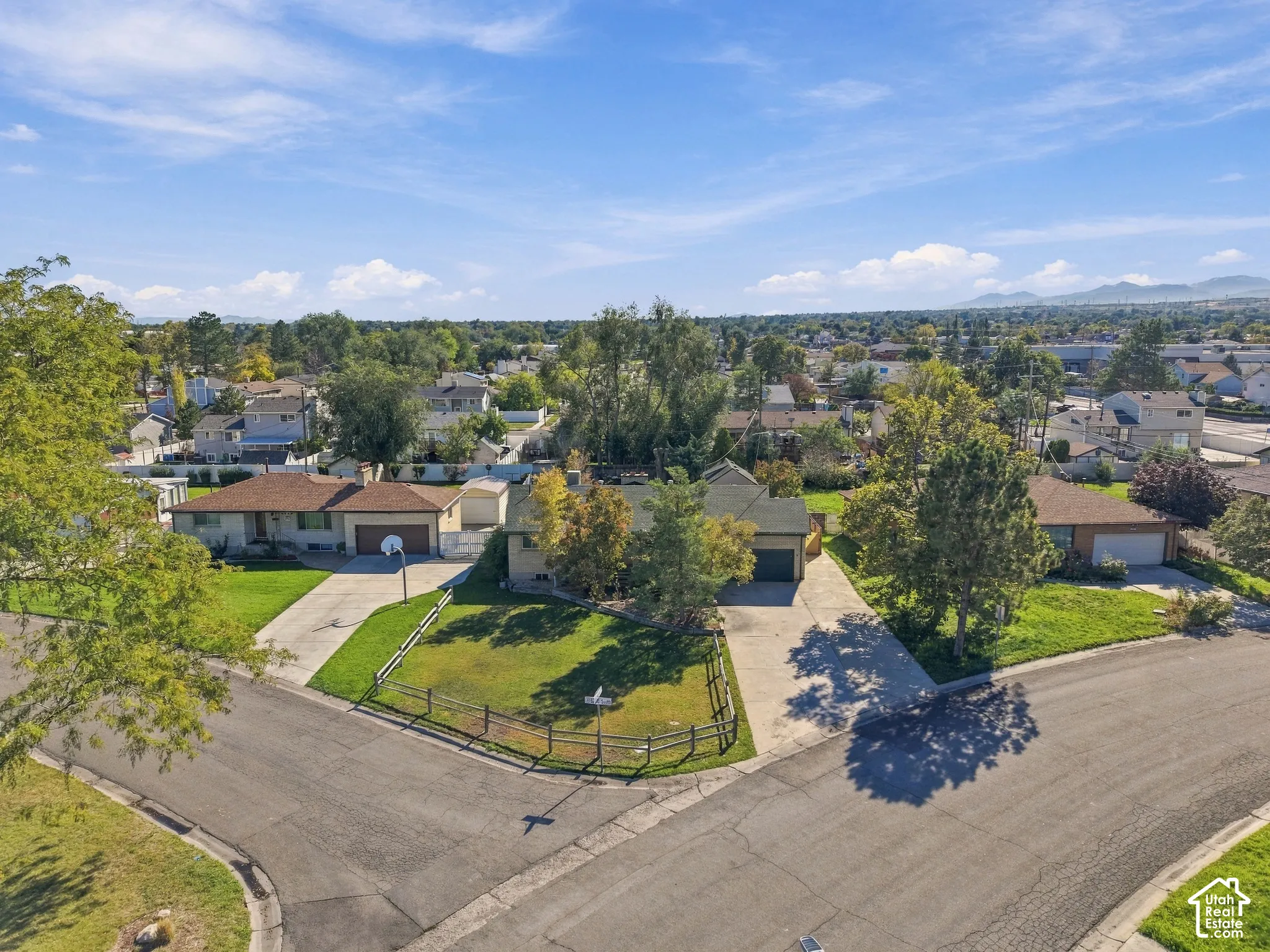 Aerial perspective featuring a corner lot, expansive front lawn, mature trees, split-rail wood fencing, and multi-car concrete driveways