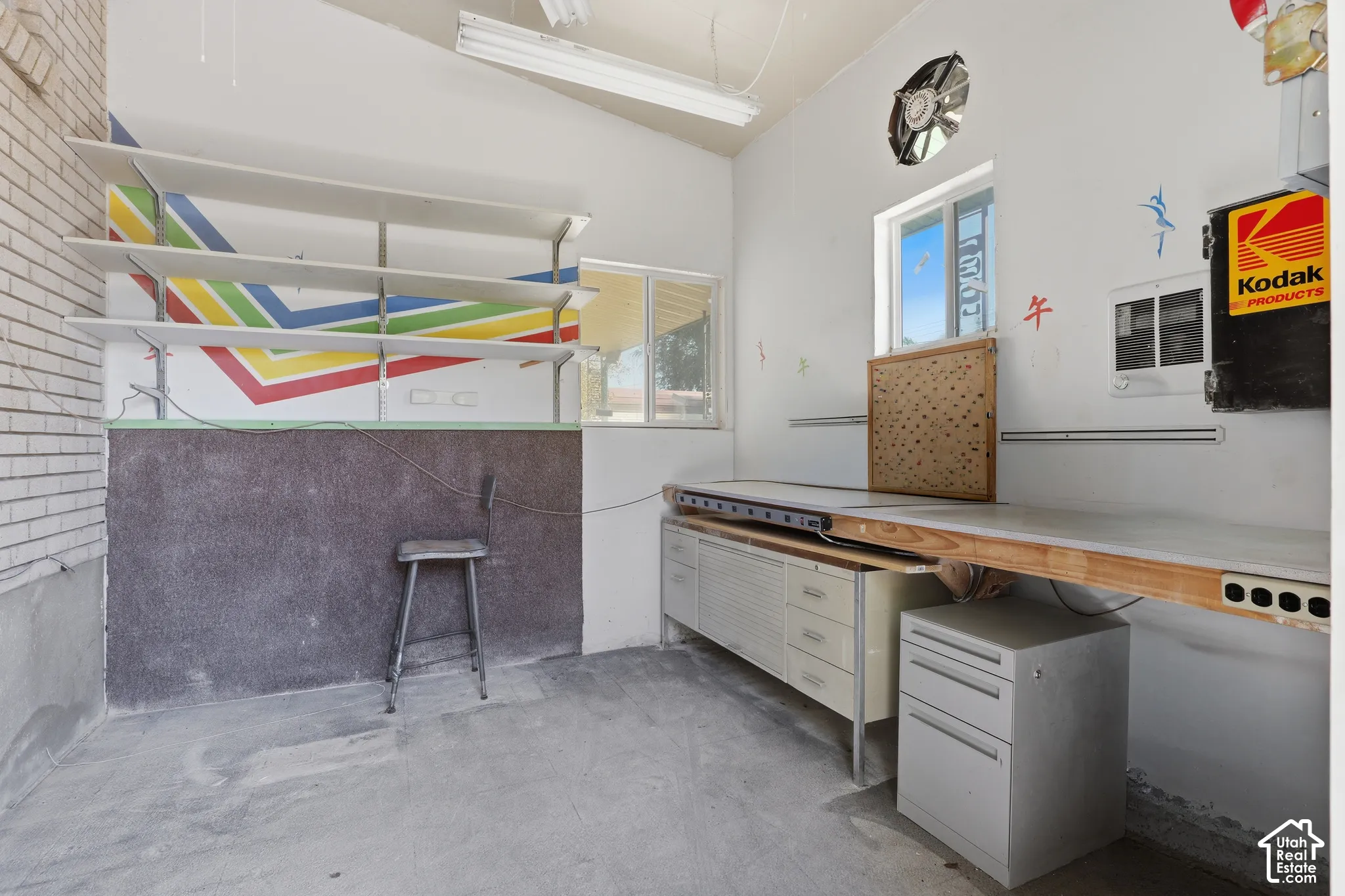 Workspace featuring built-in multi-tier wall shelving, expansive fixed workbench, integrated metal cabinetry, concrete flooring, and an exhaust fan
