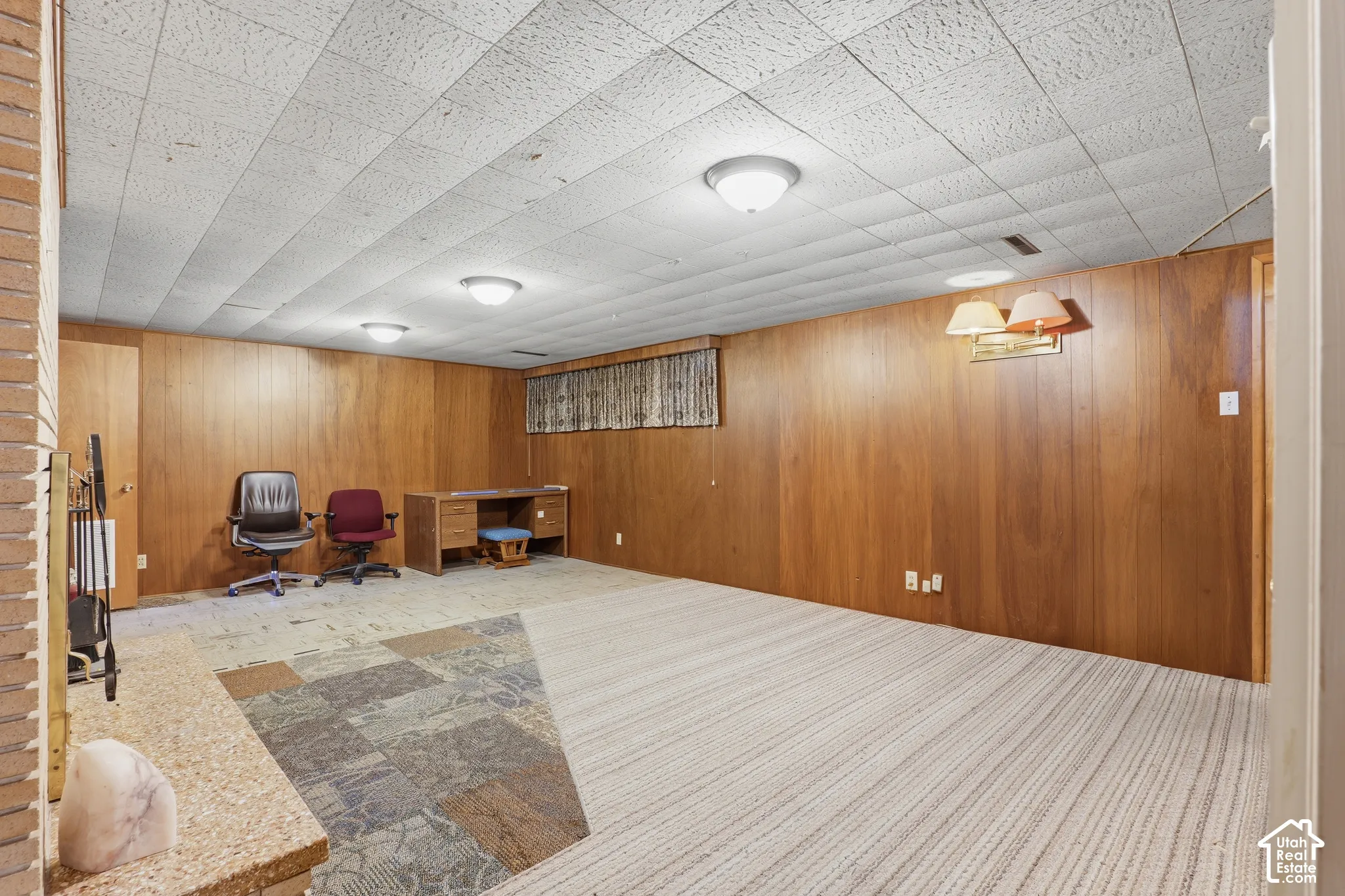 Wood-paneled walls, drop ceiling, multi-tone patterned carpeting, and masonry hearth
