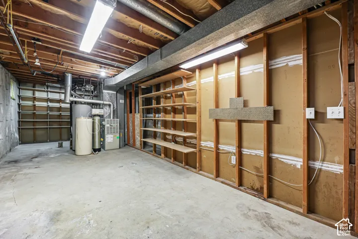 Unfinished utility area featuring exposed wood stud framing, concrete flooring, built-in wooden wall shelving, and overhead linear fluorescent lighting