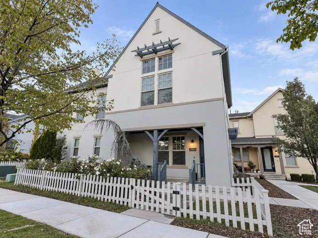 Two-story exterior featuring a white stucco facade, decorative upper-level timber pergola, covered front porch with timber bracket supports, and a white picket fence