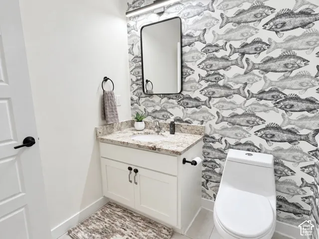 Custom vanity with stone-style countertop and undermount sink, patterned wallpaper, rectangular mirror with rounded edges, matte black hardware, and single-piece toilet