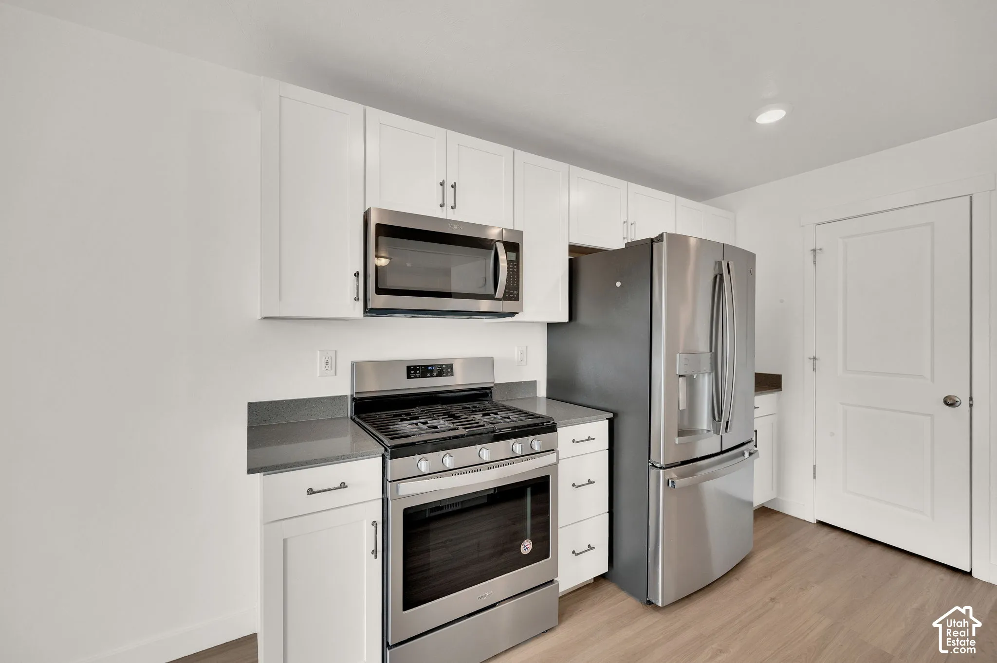 Kitchen featuring white shaker-style cabinetry, dark stone-finish countertops, stainless steel gas range with overhead microwave, stainless steel refrigerator, and wood-finish flooring
