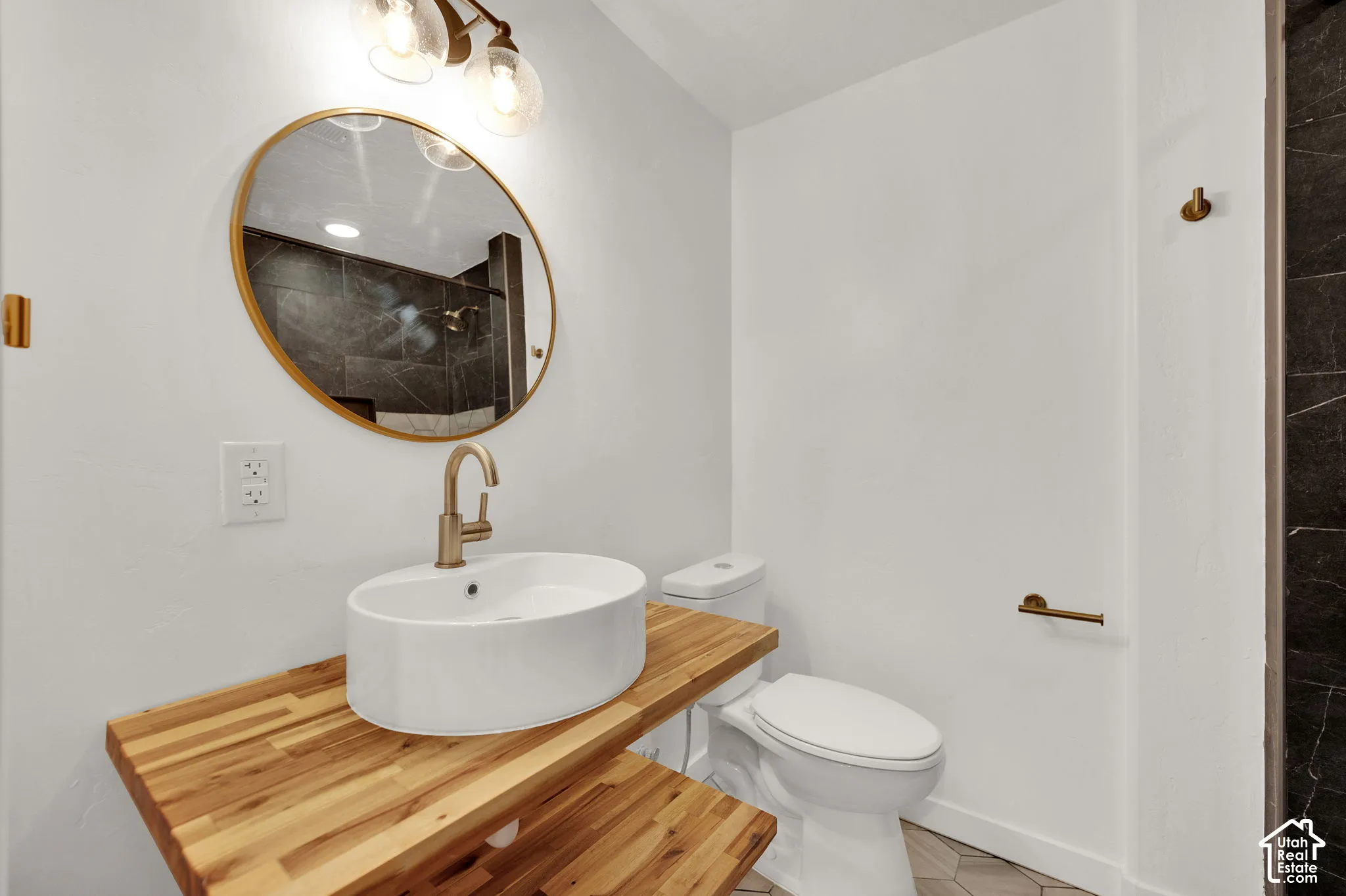 Bathroom featuring a dual-level butcher block vanity, white vessel sink with a brass-finish faucet, oval gold-rimmed mirror, and light fixture with clear glass shades