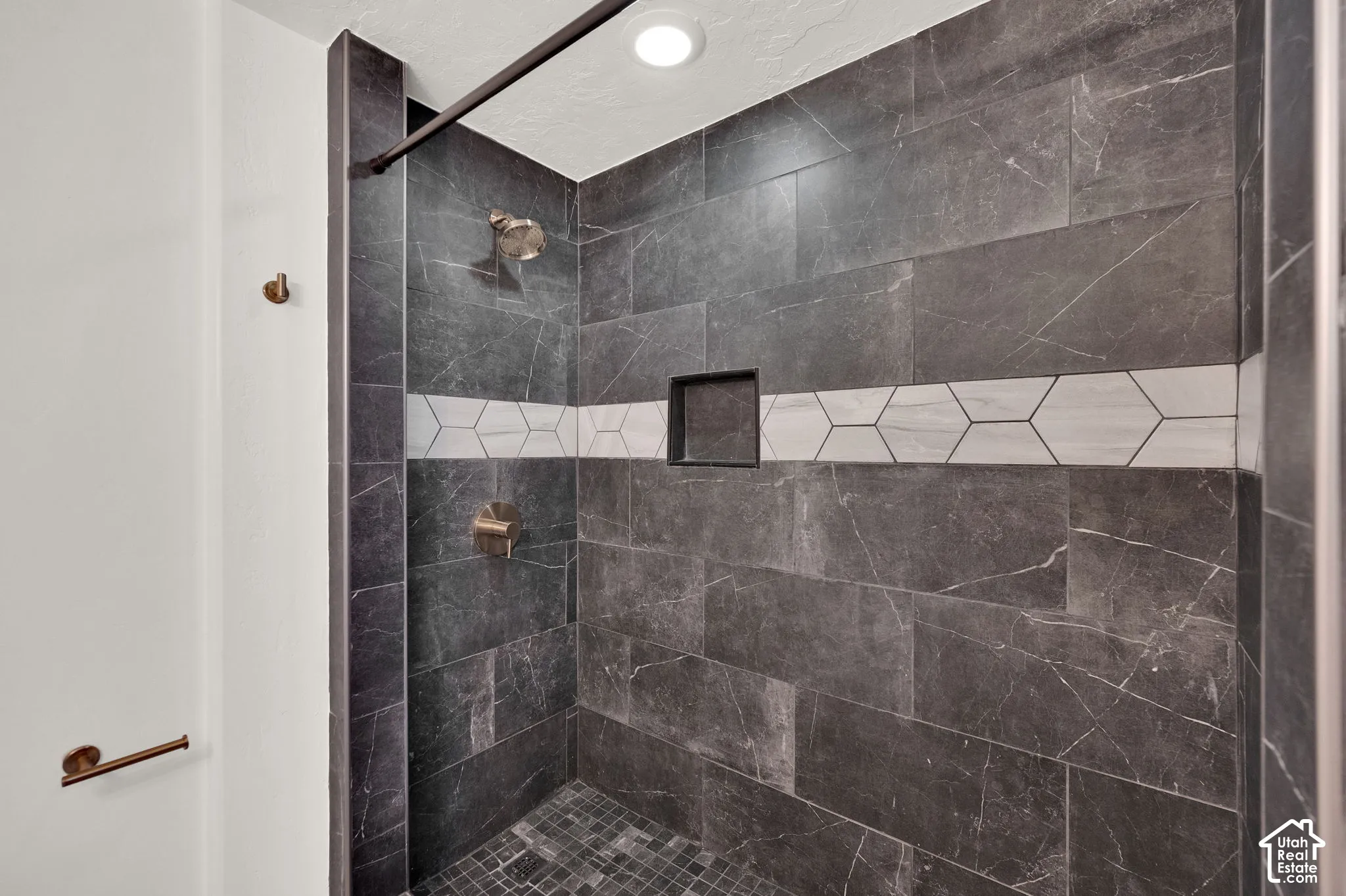 Shower stall featuring large-format dark charcoal tile, a contrasting white hexagonal tile accent band, integrated recessed storage niche, pebble-style floor tiling, and a metallic shower fixture