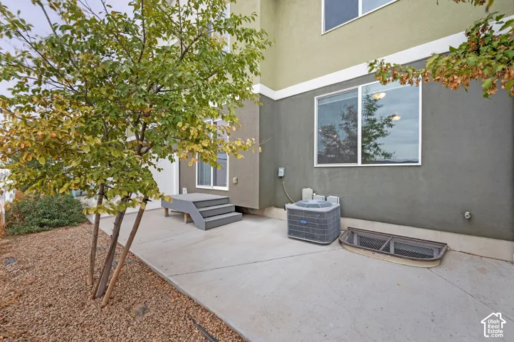 Concrete patio featuring a set of stairs, exterior HVAC unit, window well with metal grate, and multi-toned stucco siding