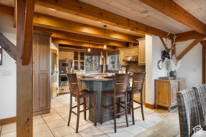 Kitchen featuring exposed timber ceiling beams, a central island with stone-surface countertop, custom wood cabinetry, stainless steel built-in double ovens, and tile flooring