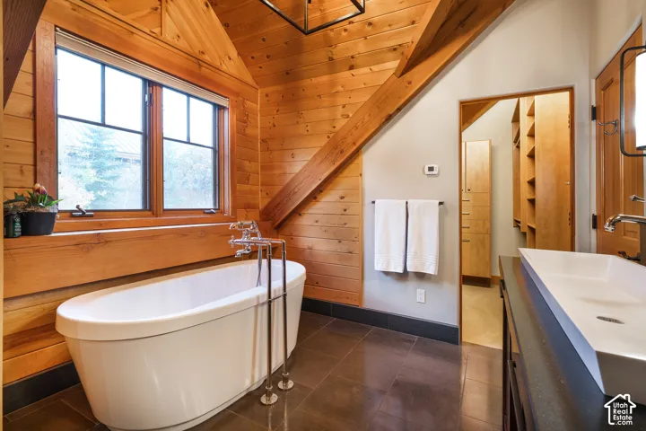 Vaulted ceiling with exposed wood beams, wood-plank wall paneling, freestanding soaking tub with floor-mounted faucet, rectangular vessel sink, and built-in shelving