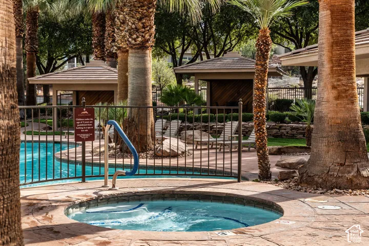 Resort-style backyard oasis featuring a sparkling spa with integrated jets, resort-gated pool access, mature palm trees providing natural shade, and expansive travertine pavers throughout the entertaining area
