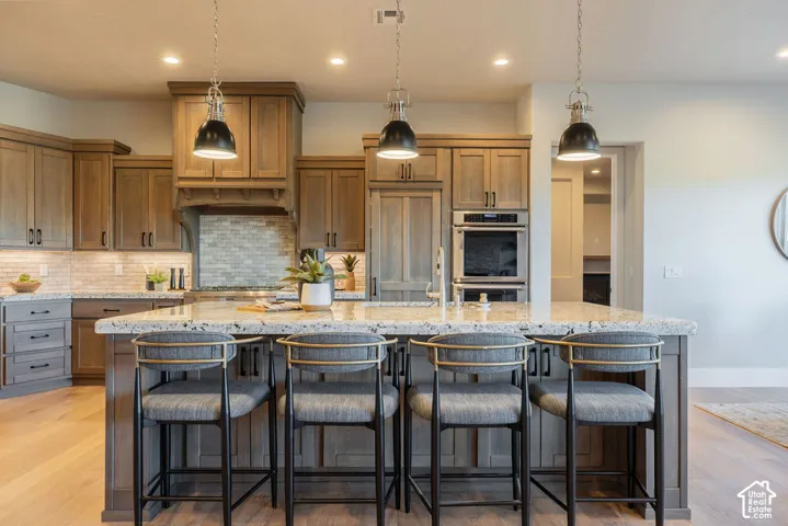 Kitchen featuring wood cabinetry, expansive stone countertop island, stone tile backsplash, stainless steel double wall ovens, and three industrial-style pendant light fixtures