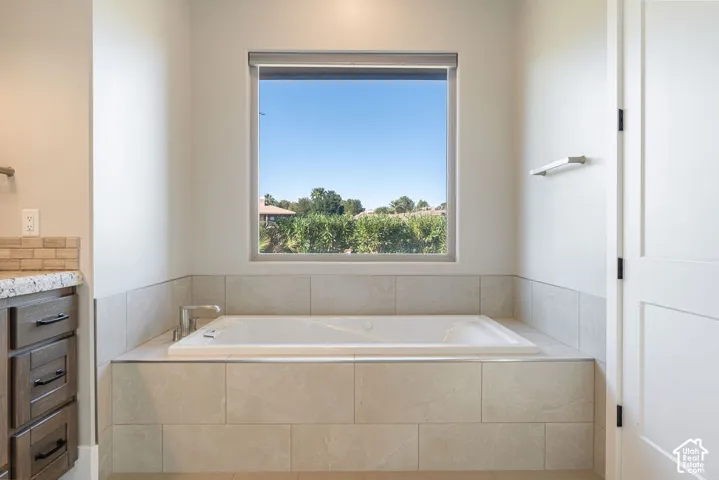 Soaking tub with tiled surround, wood-finish vanity with stone countertop, and subway tile backsplash